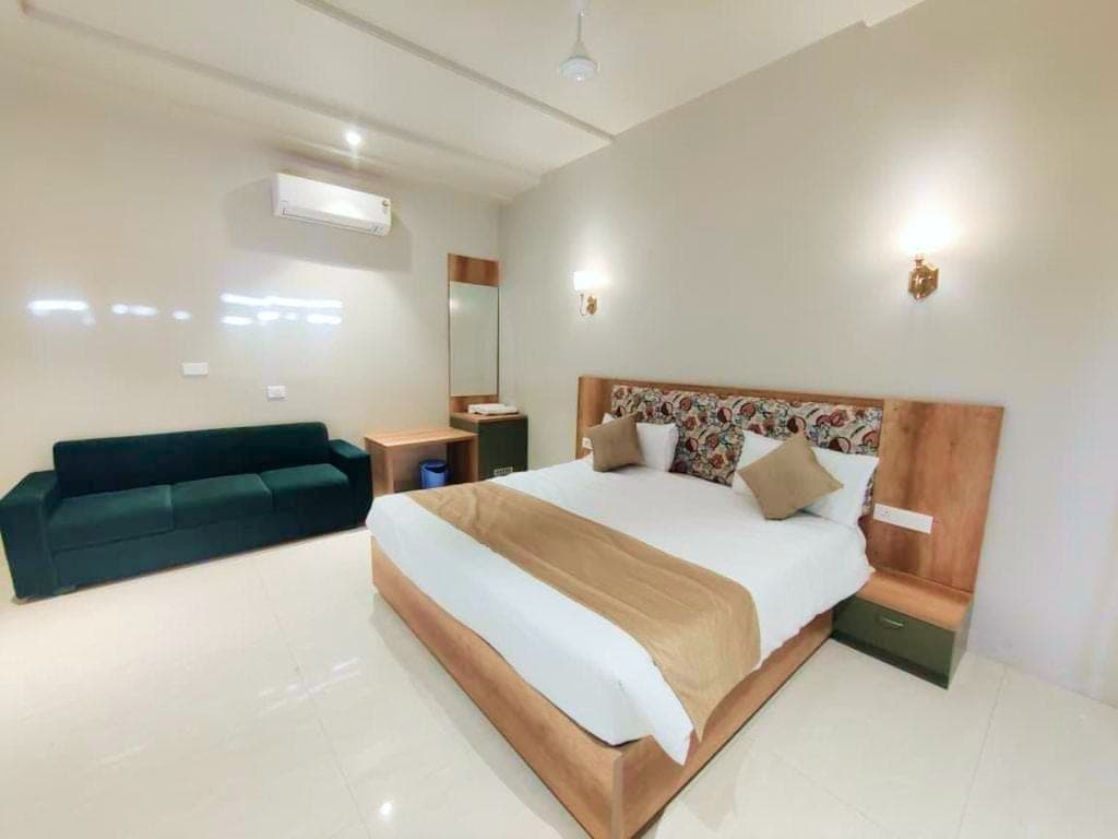 A Deluxe Room: a wide bed against a full-height wooden headboard with block-printed bolster cushions, a deep green sofa along the opposite wall, brass wall sconces and a ceiling-height air conditioner.