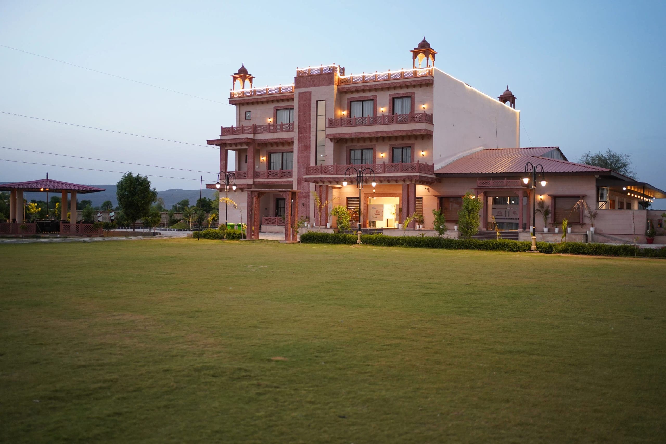 The resort at dusk from across the lawn, every balcony and parapet picked out in warm light against a deep blue sky, with the low hills of the Aravallis on the horizon behind it.
