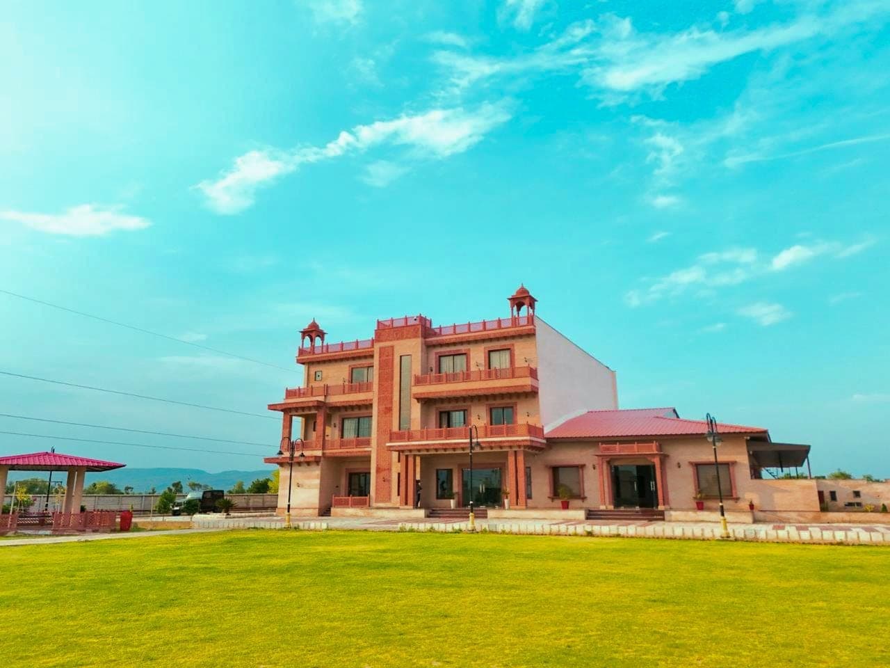 The pink sandstone main building seen across the open lawn, its two domed rooftop chhatri pavilions and tiered balconies rising against a bright sky.