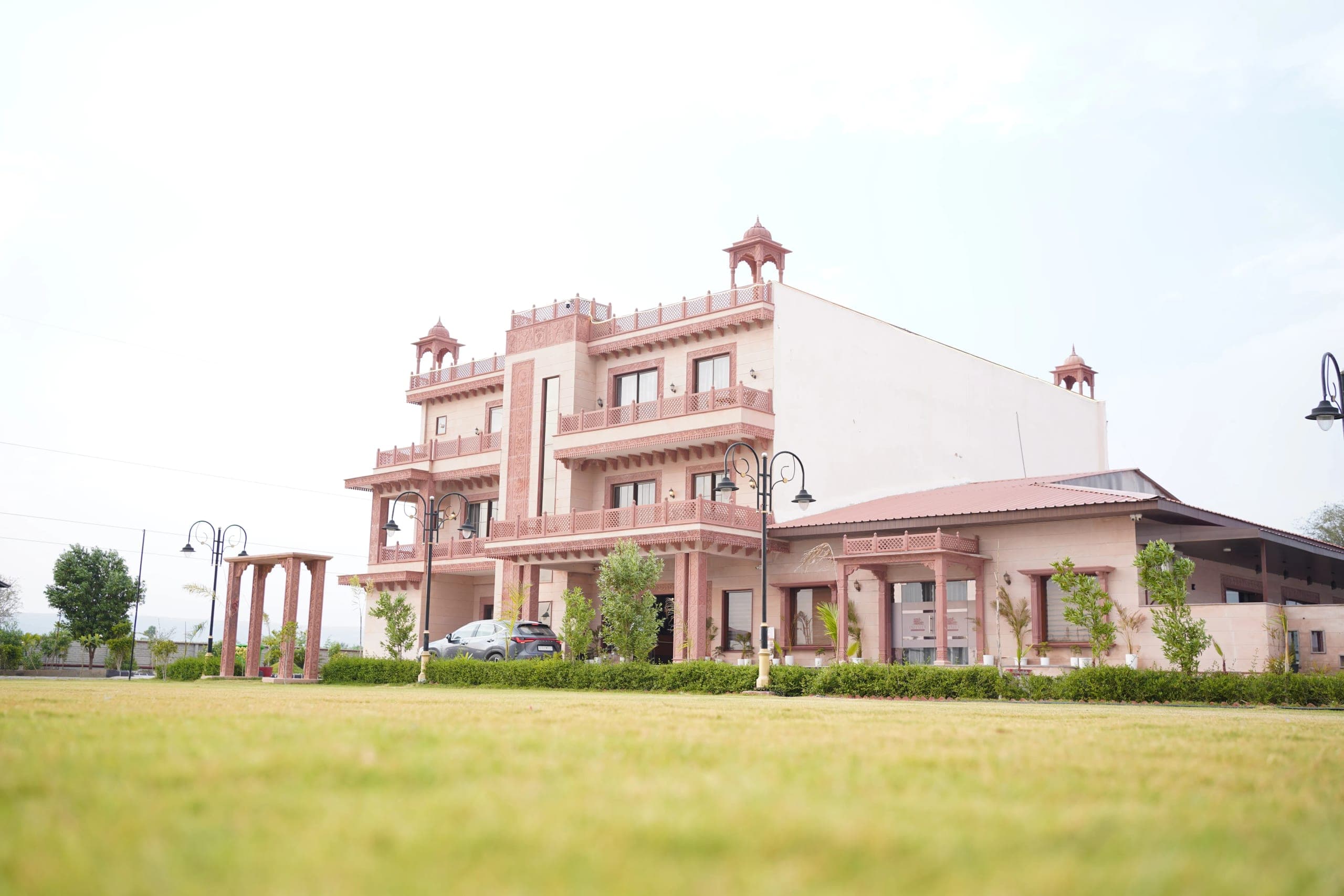 The resort seen across the lawn by day: three storeys of pink sandstone with tiered balconies and domed rooftop chhatri pavilions, a covered banquet pavilion along the right, and a jaali-topped garden arch and wrought-iron lamp posts on the grass in front.