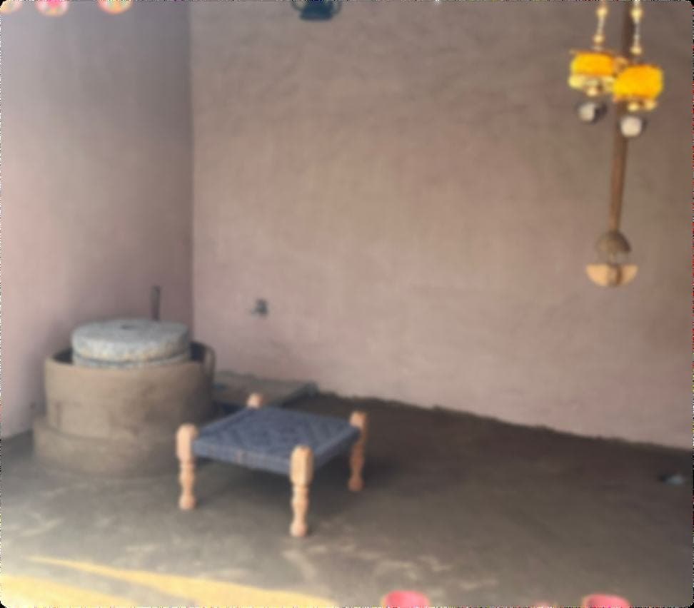 Inside the mud-walled village house: a stone hand-grinding chakki set into the corner and a low woven charpai standing on the swept earth floor.