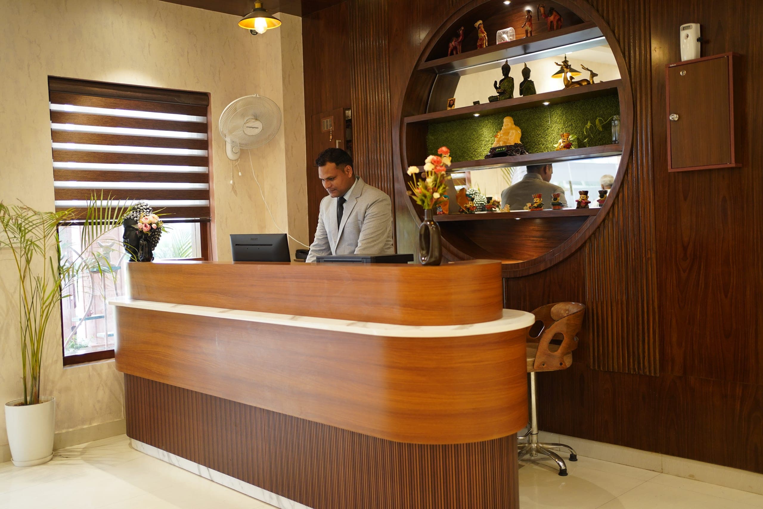 A member of the front-desk team at the curved teak reception counter, beside a tall circular display niche of backlit shelves holding carved wooden curios.