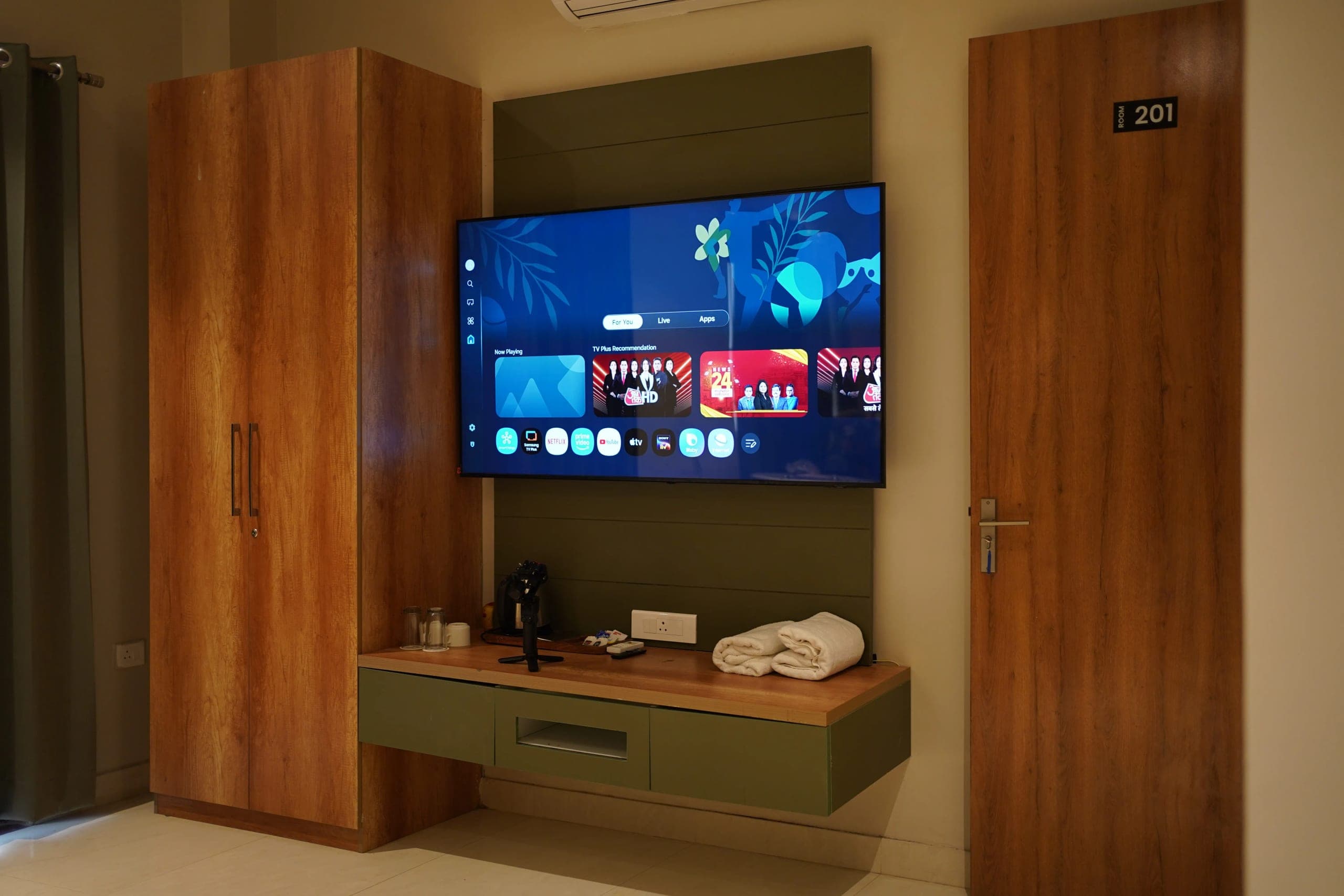 A guest room media wall: a wide flat-screen television mounted on an olive-green panel above a floating teak console holding a kettle, glasses and folded towels, with a full-height teak wardrobe to one side.