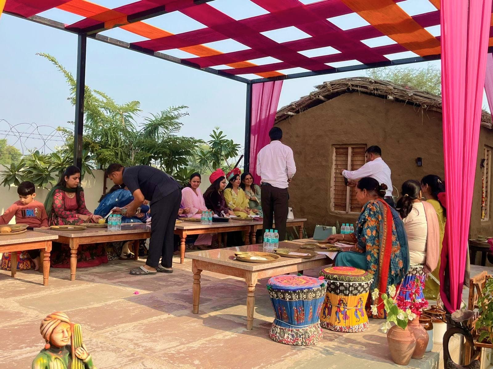 Guests eating from brass thalis at low wooden tables in the village dining courtyard, under an open canopy woven from crimson and orange fabric strips, beside a thatched mud house with a shuttered wooden window.