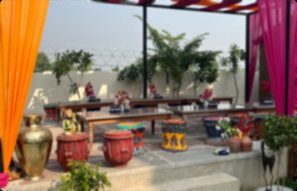 The village courtyard laid for a meal — long low wooden benches with small flower posies, framed between hanging orange and magenta drapes, with folk-painted drum stools and terracotta pots in the foreground.